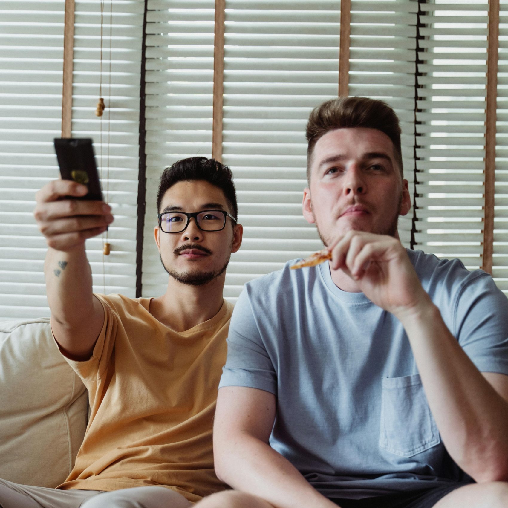 Two young men enjoying a cozy day indoors with pizza and TV, relaxing on a couch.